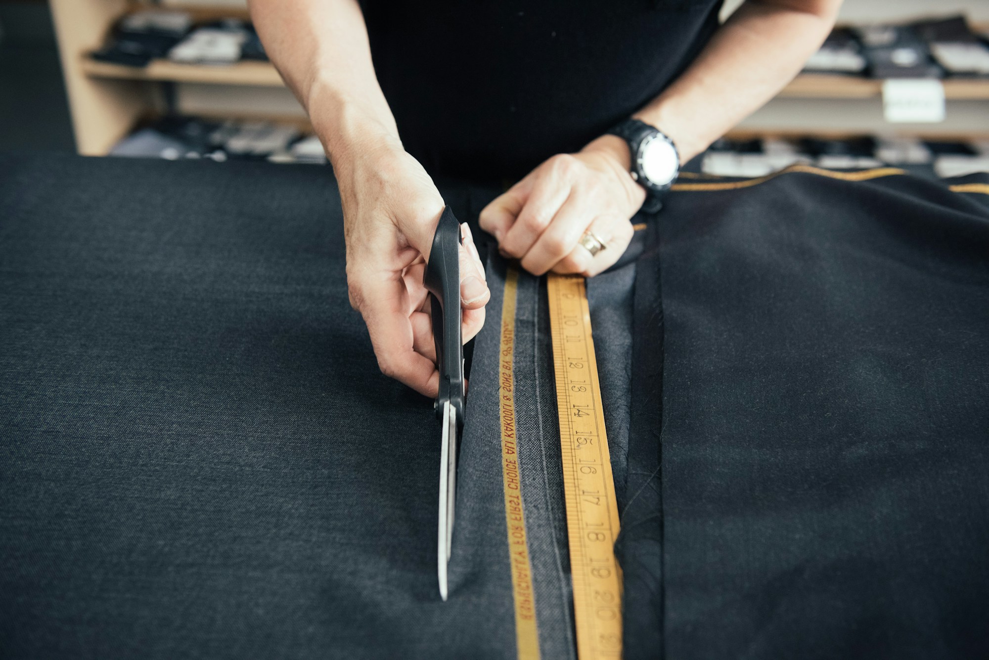 A tailor carefully cutting dark suiting fabric along a wooden ruler